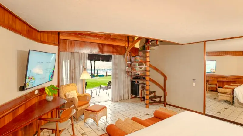Sala do bangalô com vista para o mar pela porta de vidro e escada para o mezanino