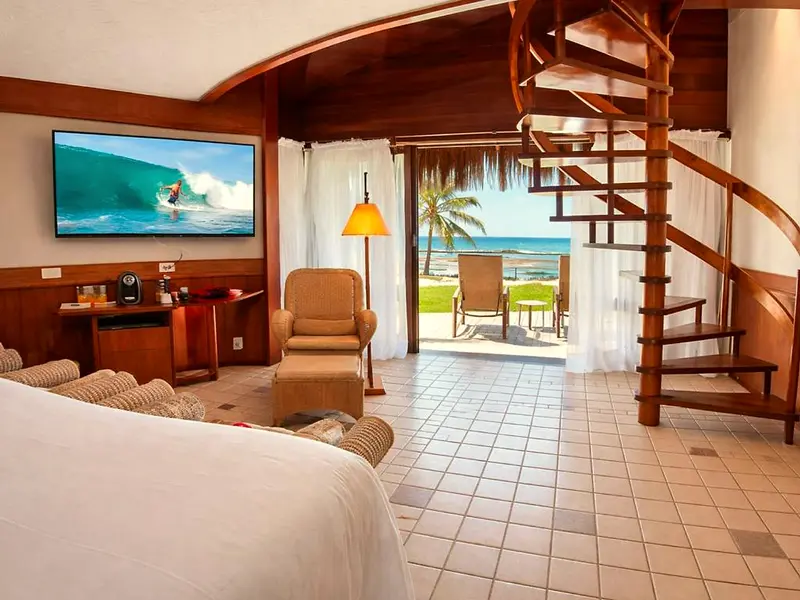 Bangalô com TV, poltronas, escada caracol e porta de vidro aberta para o mar