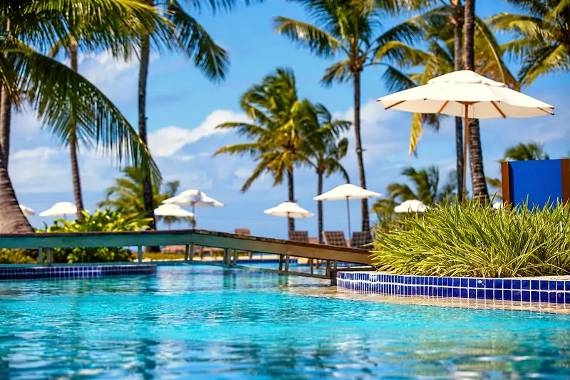 Piscina com ponte de madeira, guarda-sóis e coqueiros sob o céu azul
