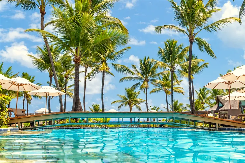 Ponte de madeira sobre a piscina, com coqueiros e guarda-sóis ao redor