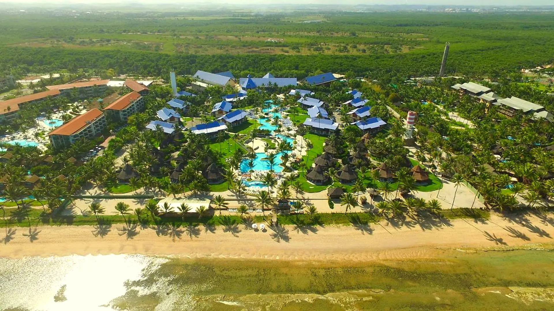 Vista aérea do resort à beira-mar, com a faixa de areia, os jardins e as piscinas entre os coqueiros